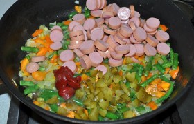 Пока овощи  пассеруются , нарезаем очищенные сосиски. Засыпаем в сковороду через 4-5 минут после фасоли, добавляем томатную пасту, приправу к мясу (или другие специи), измельченный огурчик, перемешиваем, тушим еще 3-4 минуты. 
