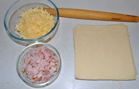 Тесто для разморозки выкладываем заранее – его можем выложить из морозилки на полку холодильника с вечера, не снимая упаковку. Натираем сыр и нарезаем ветчину.